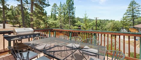 An outdoor patio with a grill, metal table, and chairs, surrounded by trees and a view of the landscape.