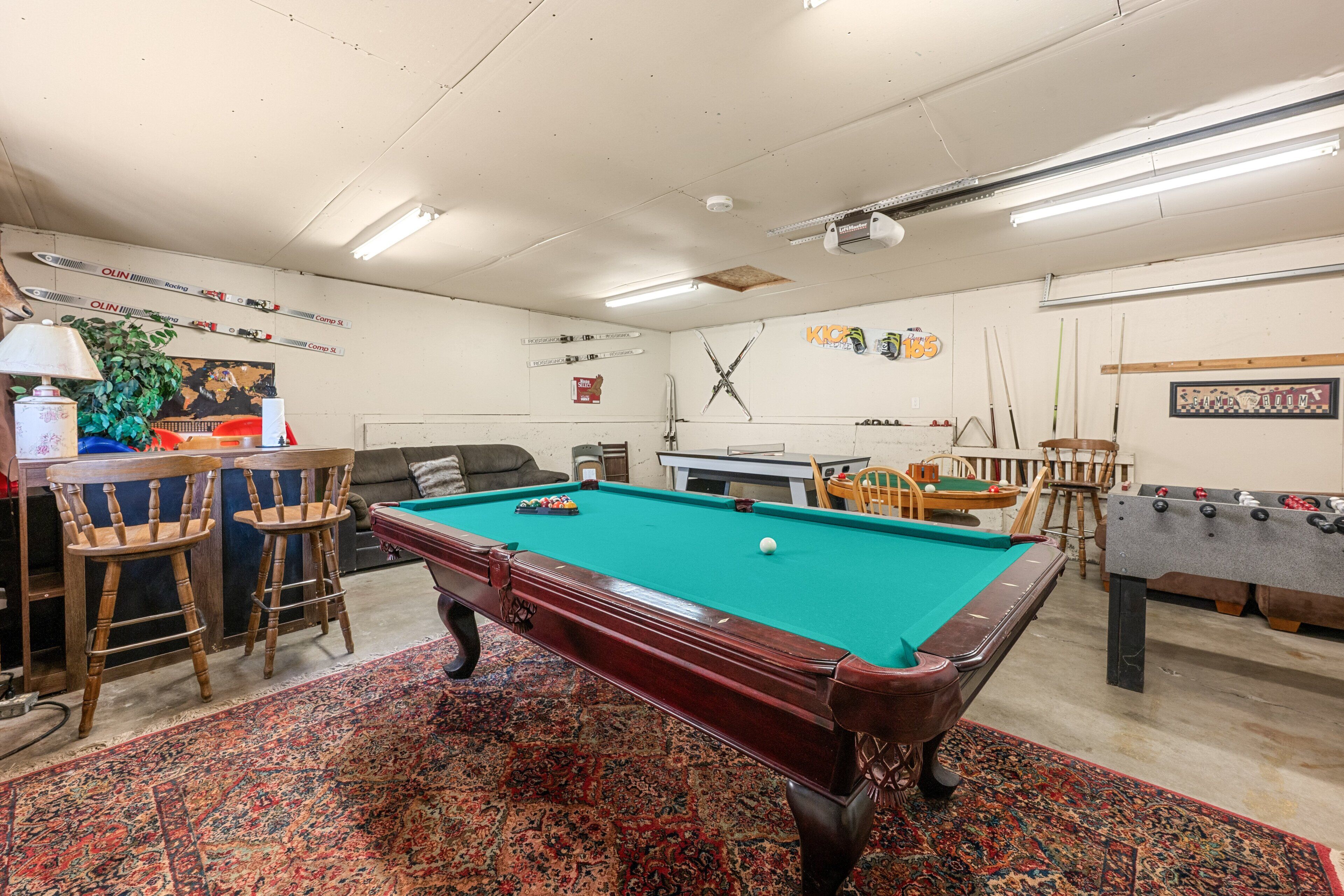 A pool room with a pool table, a foosball table, and a bar area with chairs and a lamp.