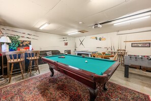 Une salle de jeux avec une table de billard, une table de baby-foot et un coin bar équipé de chaises et d’une lampe.