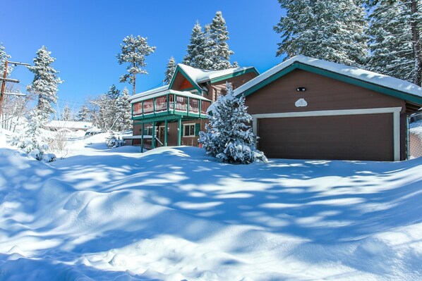 A snow-covered house with a garage, surrounded by trees and a clear blue sky.