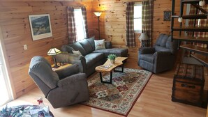 Ein gemütliches Blockhaus-Interieur mit einer Couch, Sesseln, einem kleinen Tisch mit einer Pflanze und einem Holzregal.