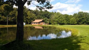 Eine Holzhütte am Seeufer mit Spiegelung, umgeben von Bäumen und einer Wiese.