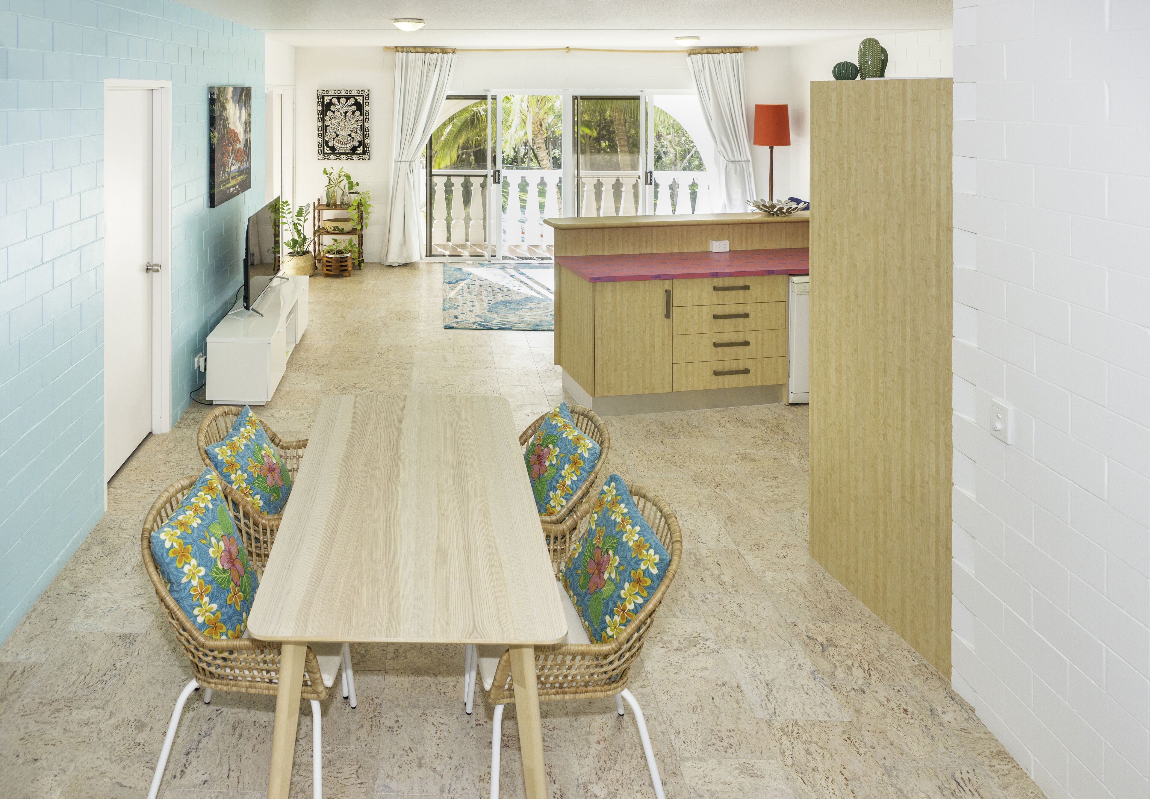 A spacious room with a wooden dining table, wicker chairs with floral cushions, a small kitchen area with a wooden counter, and a view of a balcony with white curtains.