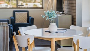Una habitación moderna con una mesa redonda, sillas blancas, un jarrón con flores azules, una computadora portátil y un televisor sobre un soporte.