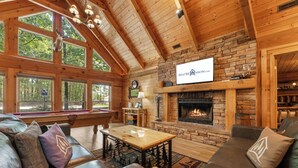 A cozy cabin interior with a fireplace, wooden walls, and a large window overlooking a forest. - Luxury Log Cabin Near Thunder Ridge_Pvt Fenced Yard & HotTub_Billiards_EV Car Charger_Free Tickets (Ridgedale)