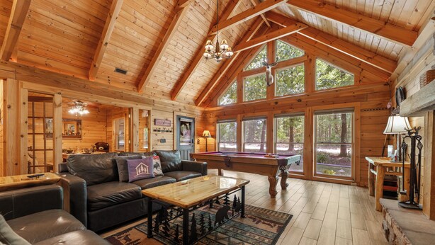 A spacious wooden cabin with a high ceiling, a pool table, a leather sofa, and large windows offering views of the surrounding forest. - Luxury Log Cabin Near Thunder Ridge_Pvt Fenced Yard & HotTub_Billiards_EV Car Charger_Free Tickets (Ridgedale)