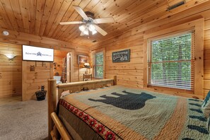 A wooden cabin interior with a bed, a ceiling fan, a sign reading 'Welcome to the Cabin', and a window with blinds.