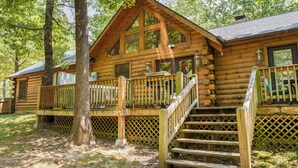 A log cabin with a wooden deck, a staircase, and a large window. - Luxury Log Cabin Near Thunder Ridge_Pvt Fenced Yard & HotTub_Billiards_EV Car Charger_Free Tickets (Ridgedale)