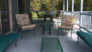 A covered porch with green metal chairs and a table, surrounded by a white fence and trees.