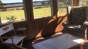 A room with a wooden interior, a chair, a table, and large windows offering a view of a grassy field and a bridge. - Ocean view, FREE BEACH PASS. Short drive to multiple beaches and water park! (Fairhaven)