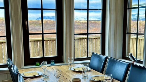 Une table à manger dressée avec des assiettes, des verres et des bols, offrant une vue sur un paysage montagneux à travers de grandes fenêtres.