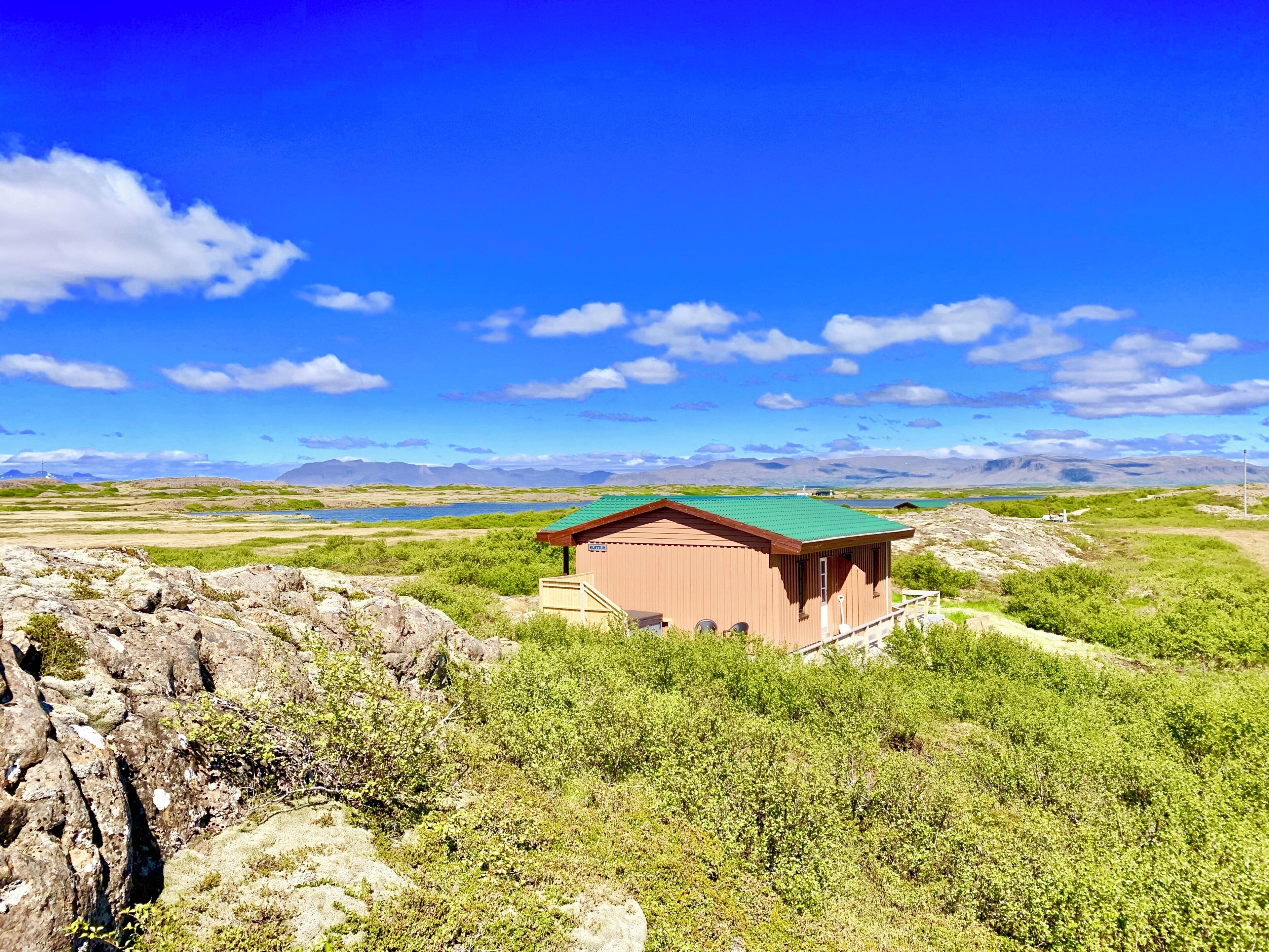 Um pequeno edifício térreo com telhado verde, cercado por terreno rochoso e vegetação rala, com vista para um corpo d'água e montanhas distantes.
