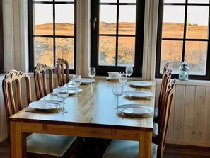 Une table à manger dressée pour six personnes, avec des chaises, de grandes fenêtres donnant sur un champ et une lanterne posée sur le rebord de la fenêtre.
