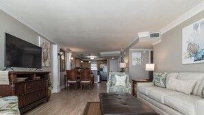 A spacious living room with a flat-screen TV, a leather ottoman, and a white sofa. There is a dining area with wooden chairs and a kitchen in the background. - BEACHSIDE 2 SANDESTIN SUMMER DISCOUNTS! BEACH FRONT 1 BR NEW POOL & HOT TUB! (Miramar Beach)