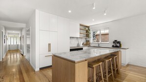 A modern kitchen with white cabinetry, a large island with stools, and a view of a brick wall through a window. - Pet friendly Riverside Retreat (Barwon Heads)