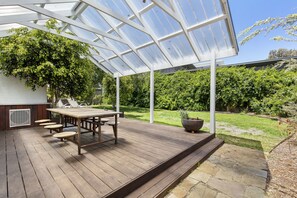 A covered patio with a wooden table and chairs, a potted plant, and a view of a grassy garden.