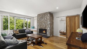 A modern living room with a stone fireplace, leather sofas, wooden flooring, and large windows offering a view of greenery. - Pet friendly Riverside Retreat (Barwon Heads)