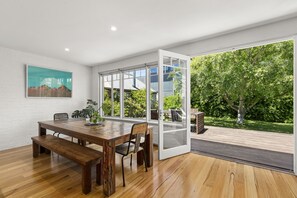 A spacious dining area with a wooden table and benches, large windows offering a view of a garden and a modern building.