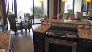 A modern kitchen with a built-in stove, a dining area with chairs and a table, and a view of the outdoors through large windows.