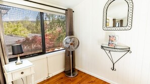 A room with a standing fan, a window with curtains, a small table with a pitcher, and a wall-mounted mirror. - ALLAMBIE COTTAGES - VILLA 2: Bright (Bright)