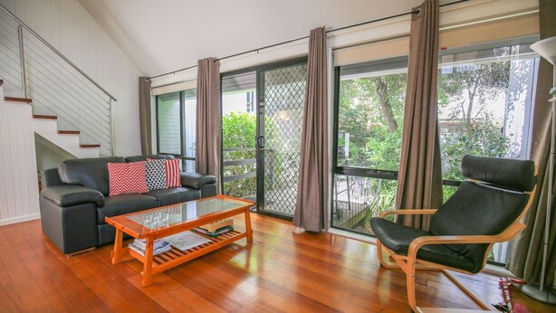 A modern living room with a sofa, a coffee table, and a chair. There is a staircase on the left and a large window with curtains on the right. - ALLAMBIE COTTAGES - VILLA 2: Bright (Bright)