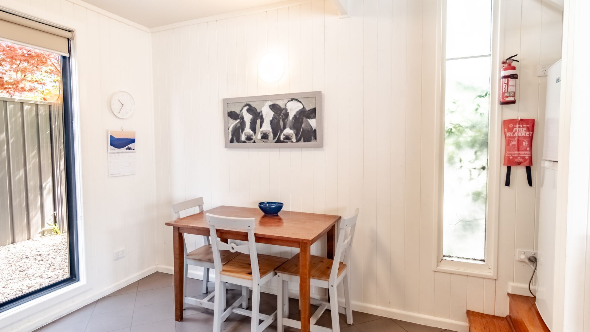 A dining area with a wooden table and white chairs, a framed picture on the wall, and a fire extinguisher mounted on the wall.