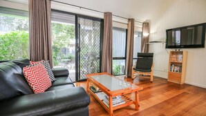 A living room with a black leather sofa, a glass coffee table, a wooden bookshelf, and a TV mounted on the wall. - ALLAMBIE COTTAGES - VILLA 2: Bright (Bright)