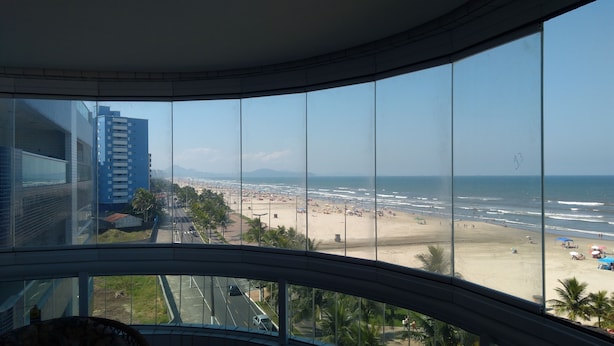Une vue depuis une fenêtre d’un immeuble de grande hauteur, donnant sur une plage, avec un ciel dégagé, des palmiers et une chaîne de montagnes lointaine.