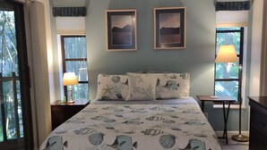 Quarto com uma cama grande, duas imagens penduradas na parede, um ventilador de teto e uma janela com persianas.
