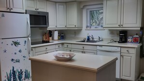 Uma cozinha moderna com armários brancos, uma ilha central, uma geladeira com adesivos decorativos, micro-ondas, lava-louças e uma janela com vista para o verde.