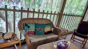 Um sofá de vime com duas almofadas, uma mesinha com uma vela e um objeto decorativo, e uma mesa redonda de madeira com um castiçal e uma caixa roxa.