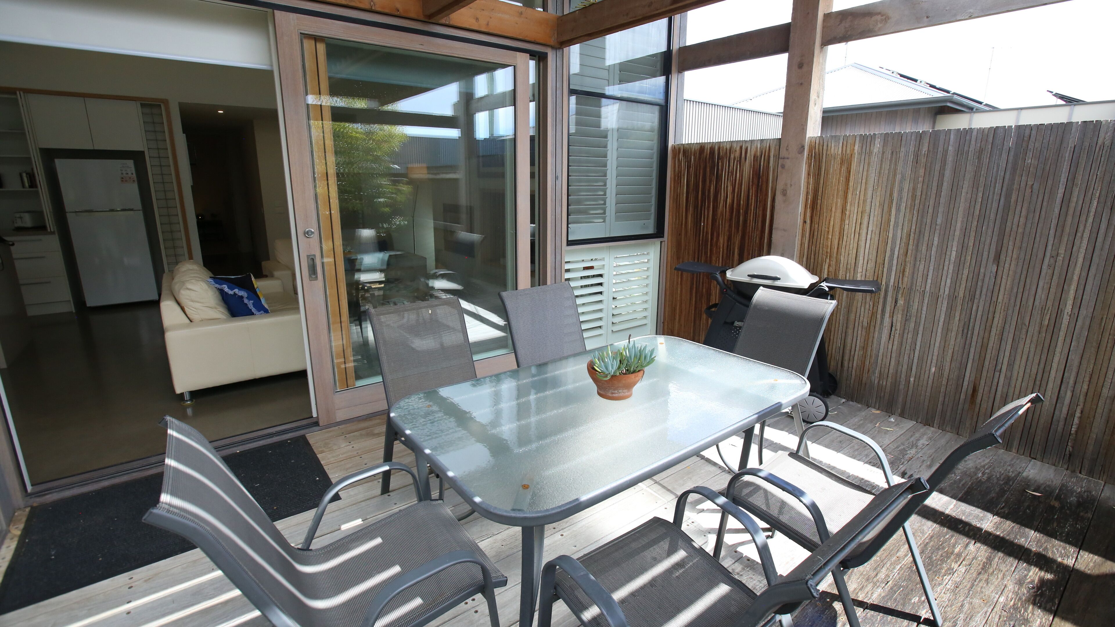 A glass-top table with chairs, a potted plant, and a sliding glass door leading to a living area with a sofa and a refrigerator.