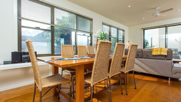 A dining area with a wooden table and woven chairs, large windows with blinds, and a ceiling fan.