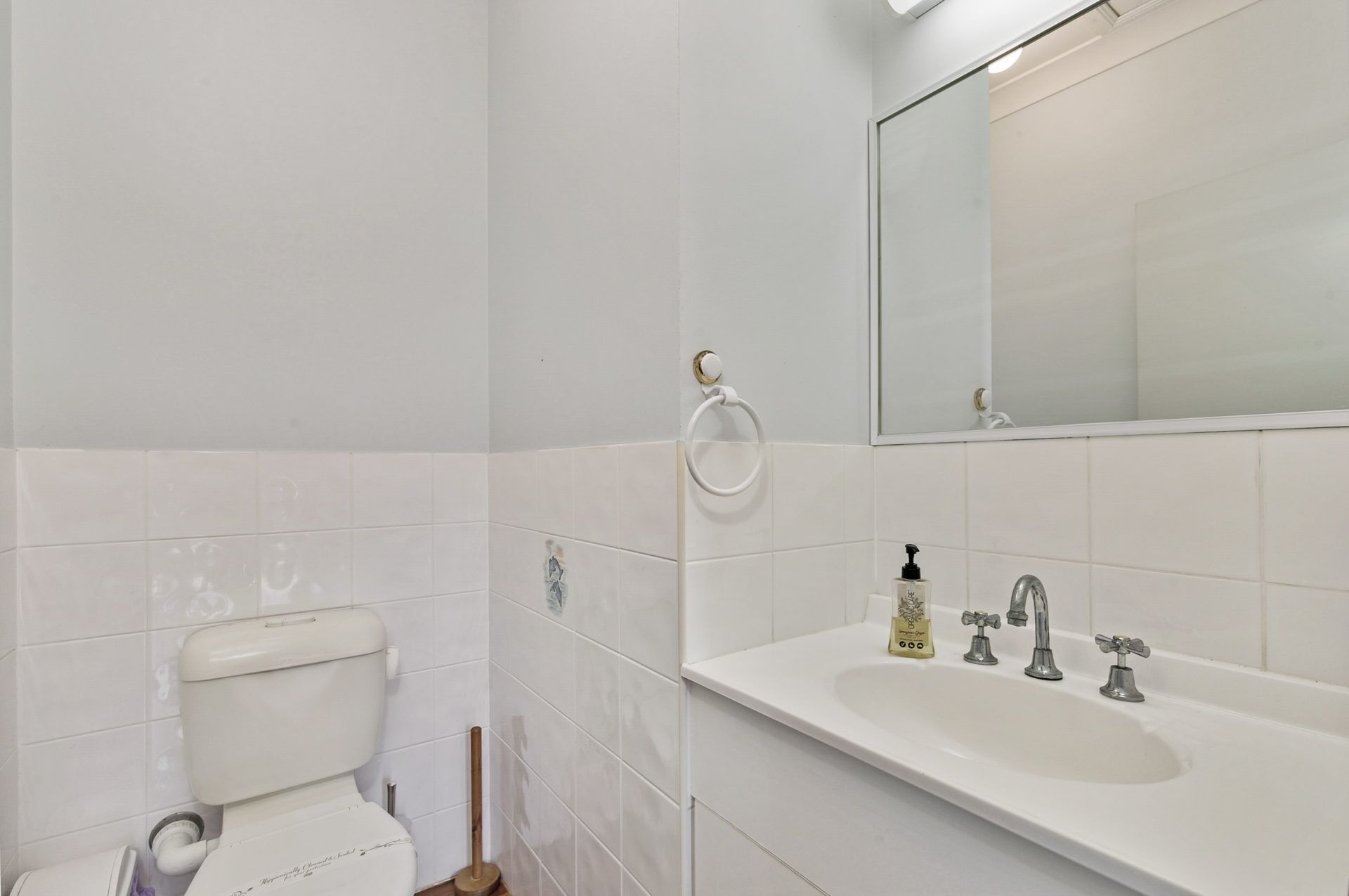 A bathroom with white tiles, a toilet, a sink with three faucets, and a mirror.