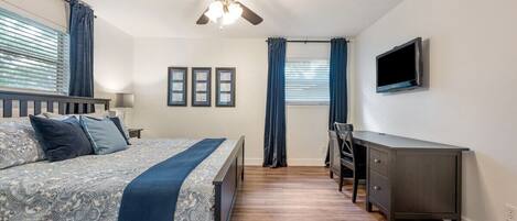 A bedroom with a bed, a desk, a chair, a ceiling fan, and a TV.