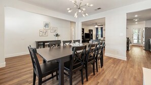 Un vaste espace à manger avec une table et des chaises en bois foncé, un lustre moderne et une vue sur la cuisine.