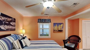 A bedroom with a bed, a ceiling fan, a window with curtains, a chair, and artwork on the walls. - Charming Tybee home w. patio, grill + pet friendly! (Tybee Island)