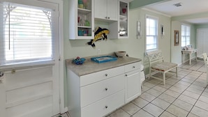 A kitchen with white cabinets, a countertop with a bowl and a blue tray, and a chair in the dining area. - Charming Tybee home w. patio, grill + pet friendly! (Tybee Island)