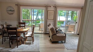 A spacious living area with a dining table, chairs, a sofa, and a large window offering a view of the outdoors. - Lakefront- Rhode Island; Indian Lake, Wakefield : 223 Indian Trail S. (Wakefield)