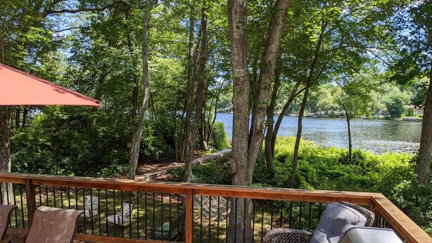 A wooden deck with outdoor furniture, a grill, and a view of a lake surrounded by trees. - Lakefront- Rhode Island; Indian Lake, Wakefield : 223 Indian Trail S. (Wakefield)