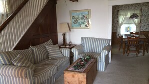 A cozy living room with striped sofas, a wooden coffee table with a floral arrangement, a painting on the wall, and a dining area with wooden furniture.