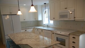 A kitchen with white cabinets, a granite countertop, a built-in microwave, and a stove. There are blue bar stools and pendant lights. - Designer Home in Mashpee with central A/C (Mashpee)