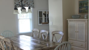 A dining area with a wooden table, six chairs, a chandelier, a window with blinds, a cabinet, and a rug. - Designer Home in Mashpee with central A/C (Mashpee)