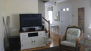 A living room with a television on a white cabinet, a wicker chair, and a staircase leading to another room. - Designer Home in Mashpee with central A/C (Mashpee)