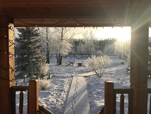 A wooden deck with snow-covered ground and trees, viewed from a porch with string lights.