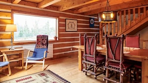 Interior de una cabaña de madera con una mesa de comedor, sillas, una lámpara, un cuadro enmarcado y una escalera.