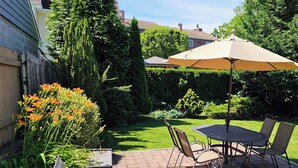 Une terrasse avec une table et des chaises, un parasol, et un jardin agrémenté de diverses plantes et d’une pelouse.