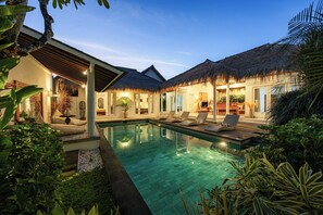A poolside area with lounge chairs, a thatched-roof structure, and a well-lit interior space.