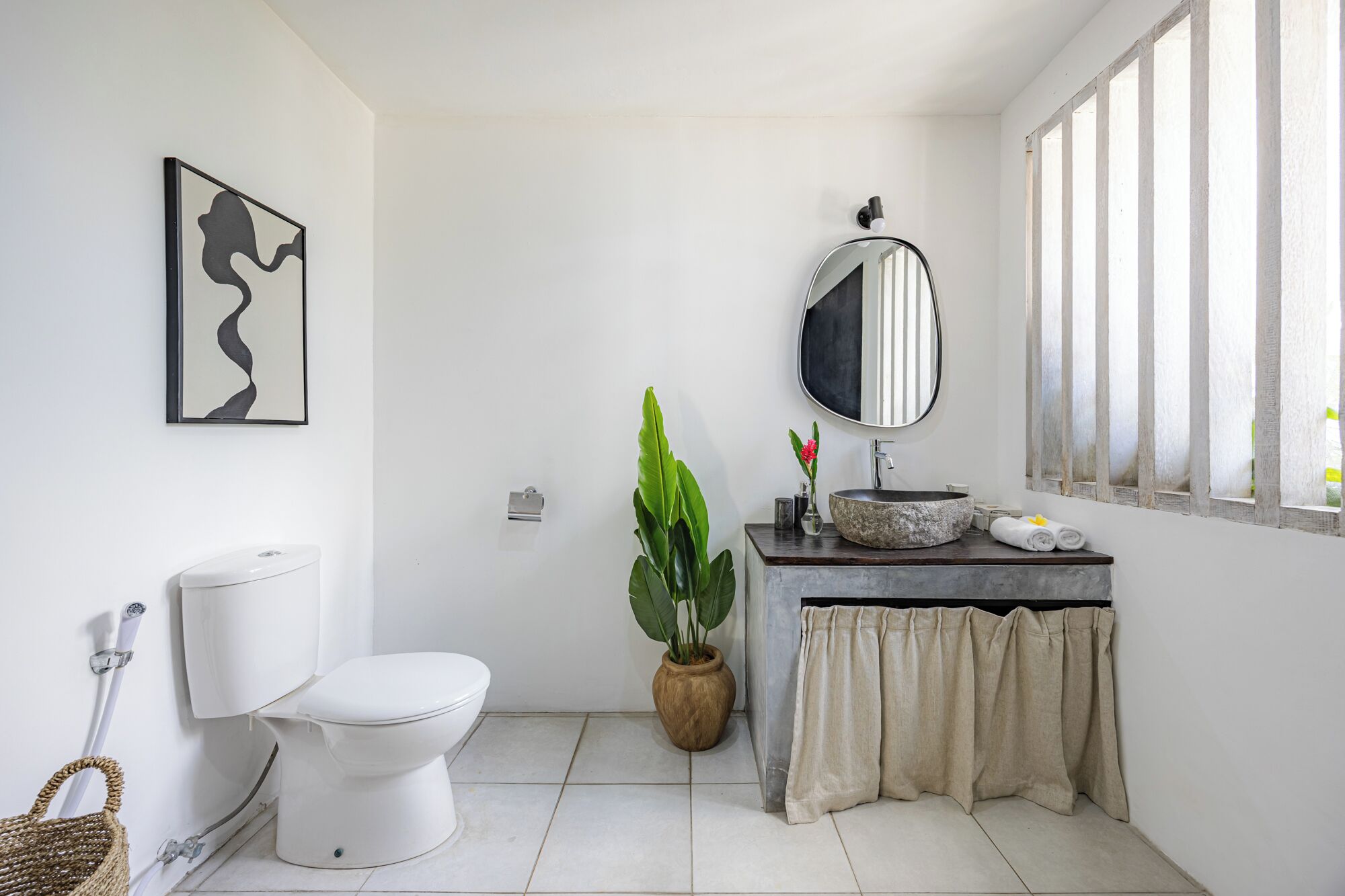 Une salle de bain avec des toilettes, un lavabo avec un miroir rond, une plante en pot et une fenêtre avec des stores verticaux.