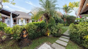 Un giardino tropicale con una statua di Buddha in pietra, una costruzione con tetto di paglia e un vialetto che conduce a un edificio bianco con porta in vetro.